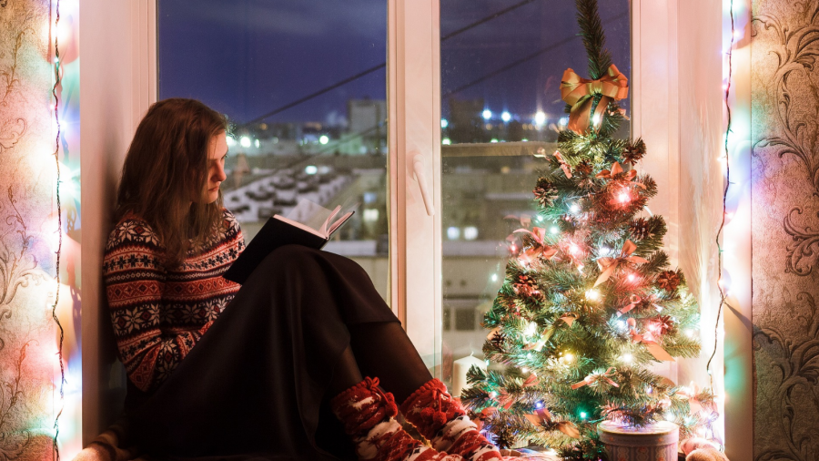 Kvinde sidder i vindueskarm med bog og lille juletræ omgivet af farverig lyskæde