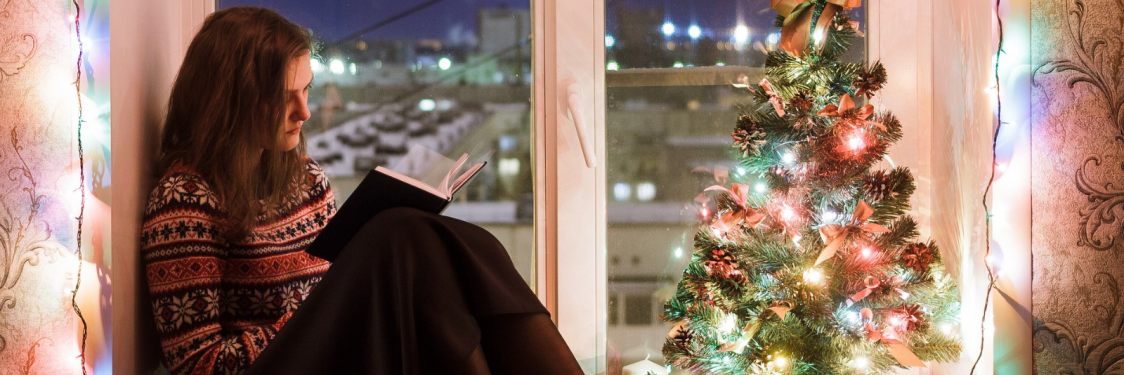 En kvinde sidder og læser i en vindueskarm ved siden af et lille juletræ med farvede lys i